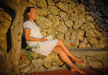 Ältere Frau sitzt mit Buch und Wein in der Nähe einer Steinmauer - DIKF00777