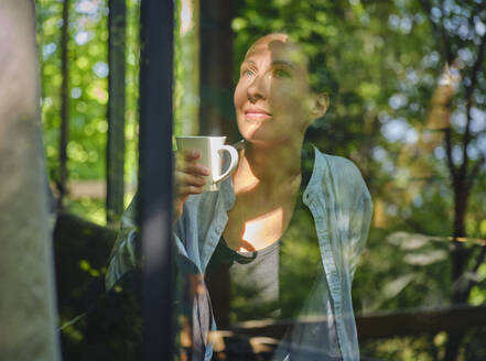 Mature woman enjoying cup of coffee and looking through window - DIKF00782