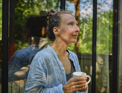 Reife Frau hält Kaffeetasse und lehnt sich ans Fenster - DIKF00783