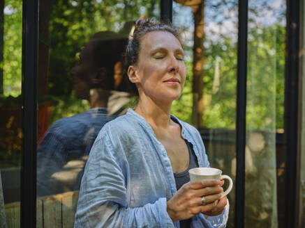 Woman with eyes closed holding coffee cup and leaning on window - DIKF00785