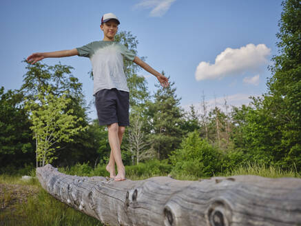 Boy with arms outstretched balancing and walking on tree trunk at forest - DIKF00788