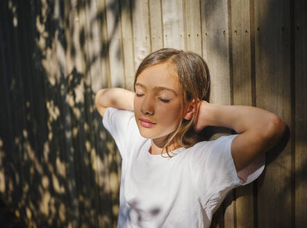 Girl with eyes closed leaning on wooden wall and enjoying sunlight - DIKF00790