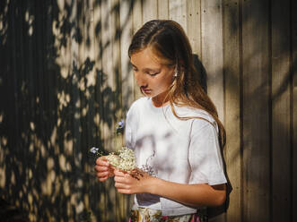 Mädchen hält Blumen und lehnt sich an eine Holzwand - DIKF00791