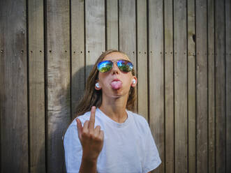 Girl wearing sunglasses and showing middle finger in front of wooden wall - DIKF00792