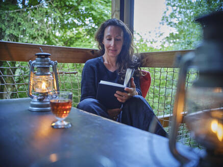 Mature woman reading book near oil lamp on porch - DIKF00795