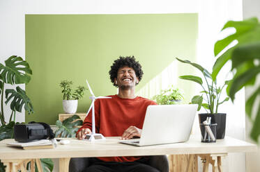 Glücklicher Freiberufler sitzt mit Laptop und Windradmodell auf dem Schreibtisch im Büro zu Hause - ALKF00816