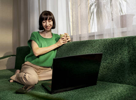 Lächelnder Freiberufler, der eine Tasse Tee in der Hand hält und sich per Videoanruf über seinen Laptop zu Hause unterhält - MBLF00142