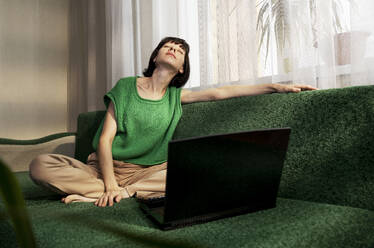 Weibliche Freiberuflerin mit geschlossenen Augen sitzt zu Hause auf dem Sofa neben dem Laptop - MBLF00143