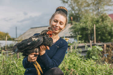 Lächelnde Frau mit Haarknoten, die einen Hahn auf einem Bauernhof hält - ADF00245