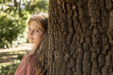 Smiling blond girl leaning on tree trunk in park - LMCF00675