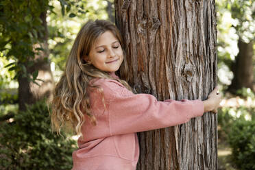 Smiling girl hugging tree in park - LMCF00679