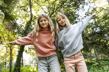 Smiling girls with arms raised standing in front of trees at park - LMCF00689