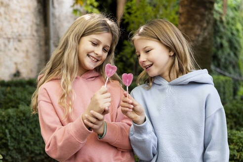 Happy girls holding heart shaped lollipop candies in park - LMCF00694