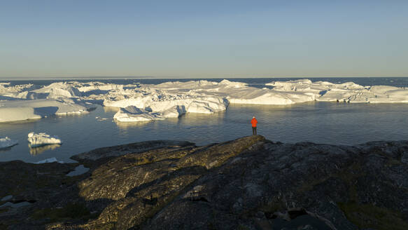 Drohnenaufnahme einer Person in Rot, die den Ilulissat-Eisfjord voller Eisberge beobachtet, Grönland, Arktis. - AAEF24189