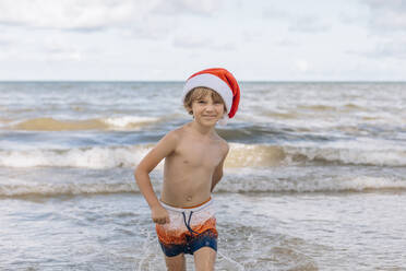 Lächelnder Junge mit Weihnachtsmannmütze läuft am Strand - EVKF00083