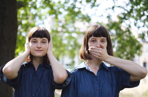 Zwillingsschwestern halten sich Mund und Ohren mit den Händen vor einem Baum zu - ASGF04763