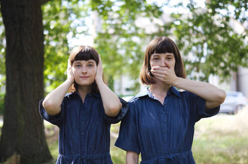 Junge Zwillingsschwestern halten sich Mund und Ohren mit den Händen vor einem Baum zu - ASGF04764