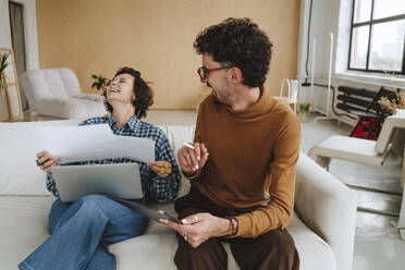 Happy graphic designer sitting with colleague laughing on sofa at office - MDOF01658