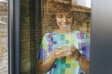 Smiling woman using smart phone seen through glass window - YTF01412