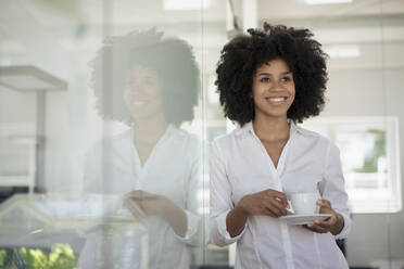 Smiling businesswoman holding tea cup leaning on glass wall at office - KNSF09972