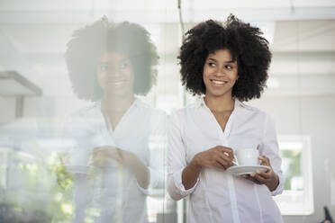 Smiling businesswoman with tea cup leaning on glass wall at office - KNSF09973