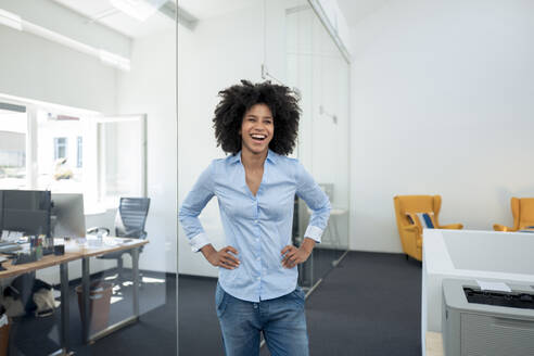 Happy businesswoman standing with arms akimbo and laughing at office - KNSF09977