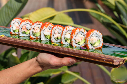 Crop unrecognizable waitress demonstrating platter with tasty California rolls covered with caviar against green plants in Asian restaurant - ADSF48827