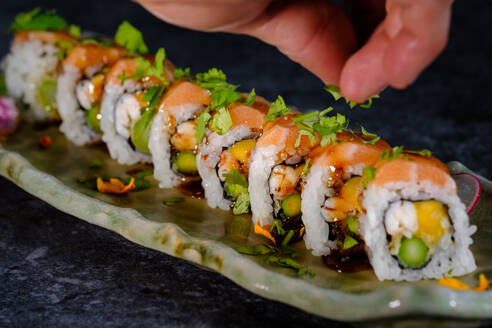 Closeup of crop anonymous person seasoning uramaki sushi with salmon and fresh vegetable with avocado served on marble table on blurred background - ADSF48829