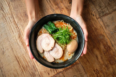 Draufsicht auf anonyme Hände einer Person, die eine Schüssel Ramen-Suppe mit Schweinefleisch, Kräutern, Gemüse und gekochtem Ei auf einem Holztisch hält - ADSF48831