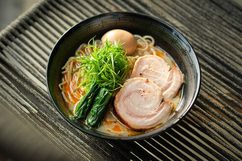 Hohe Winkel der Keramik Schüssel Ramen-Suppe mit Schweinefleisch Kräuter Gemüse und gekochtes Ei auf schwarzem Stuhl gegen unscharfen Hintergrund - ADSF48832