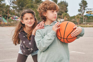 Delighted friendly teenage boy and girl with basketball standing on city street during weekend in summer - ADSF48841