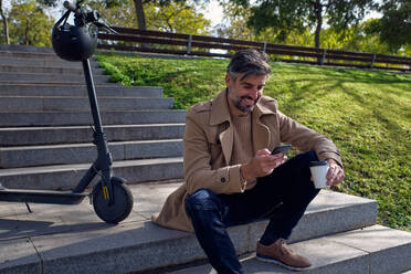 Smiling Hispanic bearded male in casual clothes sitting on stone stairs and browsing cellphone near electric scooter while having coffee break against green lawn on sunny day - ADSF48847