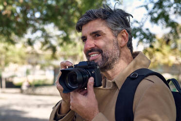 Bearded Hispanic male photographer in warm clothes with backpack taking photo on professional camera and looking away on street - ADSF48848