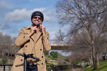 Positive active male in coat putting on protective helmet while standing on scooter in park with leafless trees on sunny spring day - ADSF48849