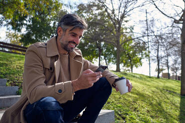 Low angle of middle aged positive unshaven Hispanic man in beige coat using cellphone and enjoying coffee break while sitting on stone stairs against trees and green hillside - ADSF48850