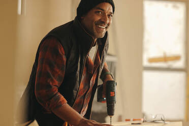 Smiling, mature contractor upgrades a kitchen during a home renovation project. Using a drill tool, the skilled carpenter works diligently to remodel the interior of the home with woodwork. - JLPSF30909