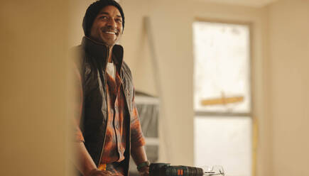 Happy contractor renovating a kitchen interior in a home. With his drill gun and tools, he upgrades the interior, bringing a smile to his face. The skilled worker's expertise shines as he remodels the house with precision. - JLPSF30910