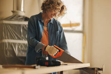 Eine glückliche Frau trägt eine Schutzbrille, während sie mit einer Kappsäge geschickt ein Holzbrett zuschneidet. Sie arbeitet an der Renovierung ihres Hauses, wobei sie sich auf die Fußleisten und die Holzverkleidung in der Küche konzentriert. - JLPSF30911