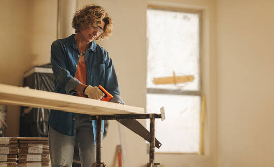 Eine Frau mit Schutzbrille arbeitet in einem Haus mit einer Kappsäge, um ein Holzbrett für ein Hausrenovierungsprojekt zuzuschneiden. Ihre geschickte Holzbearbeitung und ihr Engagement für die Verbesserung des Hauses sind in diesem Heimwerkerprojekt offensichtlich. - JLPSF30912