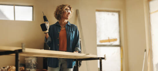 Eine glückliche kaukasische Frau renoviert die Küche ihres Hauses. Mit Handwerkzeugen und einer Schutzbrille installiert sie geschickt Fußleisten mit einer Bohrmaschine. Ihre Holzarbeiten schaffen einen einladenden und gemütlichen Innenraum. - JLPSF30913