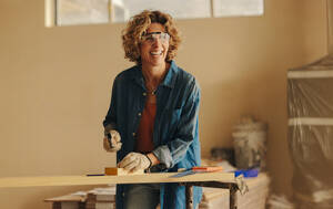 Eine glückliche kaukasische Frau renoviert ihr Haus und arbeitet an den Fußleisten in der Küche. Sie nagelt geschickt eine Holzplatte mit einem Hammer, Handwerkzeugen und einer Schutzbrille. Diese Hausrenovierung zeigt ihr Fachwissen und ihre Hingabe. - JLPSF30914