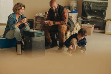 Family on a DIY home renovation project enjoys a coffee break. Parents, along with their young son, sit indoors, taking a moment to rest. Their teamwork in upgrading their house showcases dedication to creating a comfortable home. - JLPSF30922