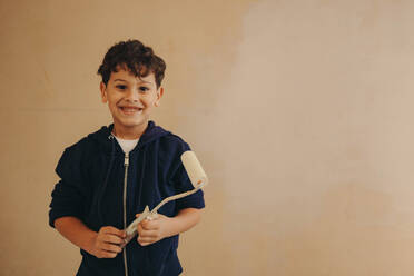 Porträt eines fröhlichen kleinen Jungen, der selbstbewusst an einer Hauswand steht und einen Pinsel in der Hand hält. Er ist ein kreativer und kluger Kopf, der sich fröhlich an der Renovierung seines Hauses beteiligt. - JLPSF30925