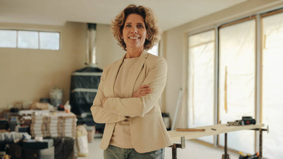 Happy, professional woman stands in a house undergoing renovation with her arms crossed. She is the project manager and homeowner, overseeing the remodeling and upgrade of the property. - JLPSF30930