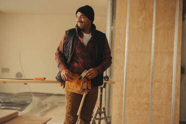 Happy male builder, wearing a tool belt, stands indoors. He is working on a home renovation, remodeling, and refurbishment project. The experienced worker's smile showcases his expertise in construction. - JLPSF30937