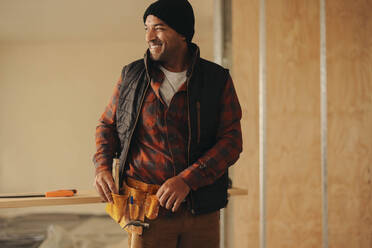 Happy, mature man with a tool belt renovates a house, improving the home for its owner. With expertise and a smile, he upgrades the interior, showcasing his skills as a DIY enthusiast. - JLPSF30938