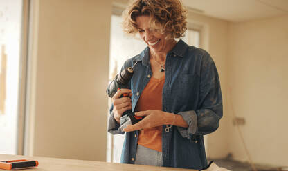 Eine Frau lächelt, während sie in einer renovierten Küche eine Bohrmaschine in der Hand hält. Bei einem Heimwerkerprojekt verschönert sie selbstbewusst ihr Haus und stellt ihre Renovierungsfähigkeiten und ihre Leidenschaft für Umgestaltungen unter Beweis. - JLPSF30942