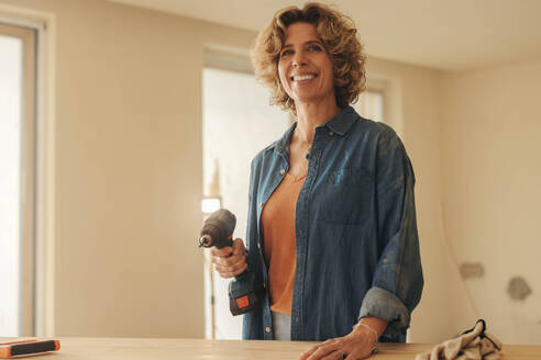 Mature, happy woman holds a power drill while working on a home renovation project in her kitchen. She stands with a smile, ready to upgrade and remodel her house with DIY expertise. - JLPSF30943