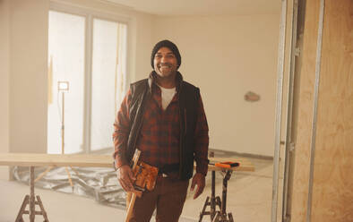 Mature male construction worker with a smile, standing in a kitchen. He is wearing a tool belt and is looking at the camera. This contractor is confident in his home renovation and remodeling skills. - JLPSF30954