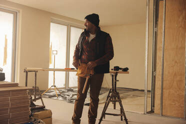 Smiling builder stands in a kitchen, wearing a tool belt, doing home renovation. He is focused on upgrading the flooring with various equipment, showcasing his expertise and dedication. - JLPSF30956